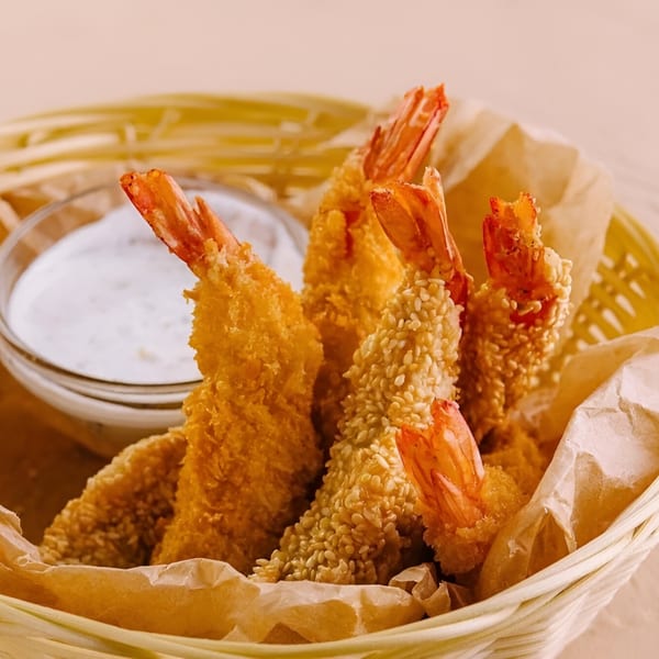 Crispy fried shrimp, some with sesame seeds, in a basket with a side of white dipping sauce.